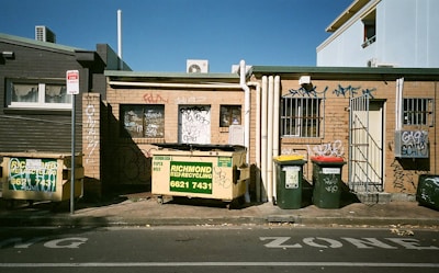 An urban alleyway featuring a small brick building with graffiti on the walls. Several recycling and trash bins are lined up against the building, with colorful graffiti marking the exterior. The area is set under a clear blue sky, creating a stark contrast between the gritty urban feel and the natural backdrop.