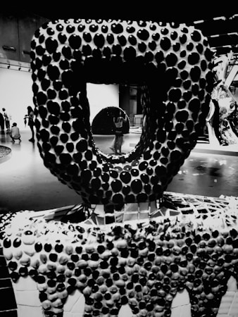 A black and white photograph featuring an abstract sculpture composed of numerous circular, bulbous shapes. The foreground displays this intricate texture, while in the background, people are seen engaging with the art exhibit. The overall aesthetic is modern and minimalistic.