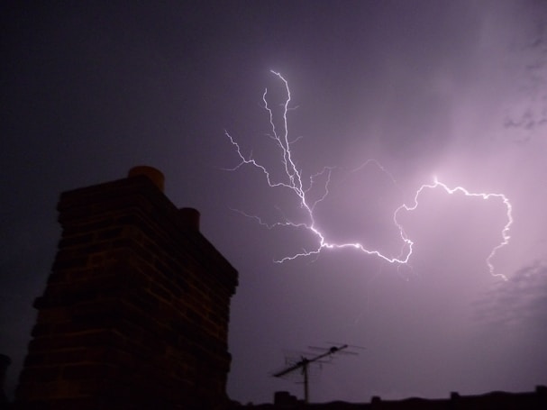 Intense lightning bolts illuminate the night sky, casting sharp contrasts against dark clouds. In the foreground, a silhouetted chimney and television antenna are visible, anchoring the dramatic scene with an urban element.