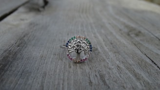 A silver ring with a tree design at the center, surrounded by small gemstones of various colors, is placed on a weathered wooden surface. The ring's intricate details and the colorful stones create a delicate and elegant appearance.