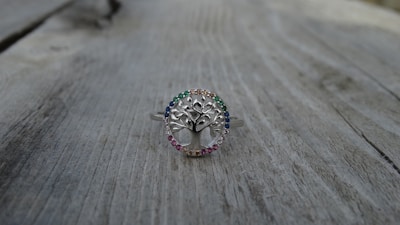 A silver ring with a tree design at the center, surrounded by small gemstones of various colors, is placed on a weathered wooden surface. The ring's intricate details and the colorful stones create a delicate and elegant appearance.