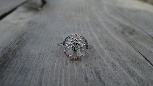 A silver ring with a tree design at the center, surrounded by small gemstones of various colors, is placed on a weathered wooden surface. The ring's intricate details and the colorful stones create a delicate and elegant appearance.