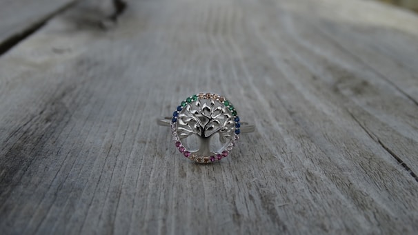 A silver ring with a tree design at the center, surrounded by small gemstones of various colors, is placed on a weathered wooden surface. The ring's intricate details and the colorful stones create a delicate and elegant appearance.