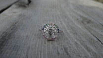 A silver ring with a tree design at the center, surrounded by small gemstones of various colors, is placed on a weathered wooden surface. The ring's intricate details and the colorful stones create a delicate and elegant appearance.
