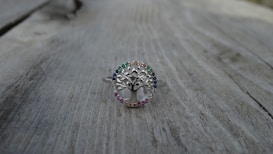 A silver ring with a tree design at the center, surrounded by small gemstones of various colors, is placed on a weathered wooden surface. The ring's intricate details and the colorful stones create a delicate and elegant appearance.