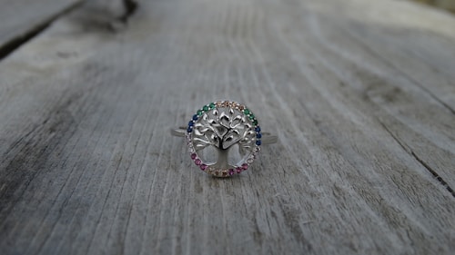 A silver ring with a tree design at the center, surrounded by small gemstones of various colors, is placed on a weathered wooden surface. The ring's intricate details and the colorful stones create a delicate and elegant appearance.