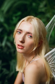 A person with long blonde hair and subtle makeup is looking towards the camera. They are adorned with sparkling decorations on the eyebrows, and they appear to have delicate fairy wings on their back. The background is filled with lush green foliage, giving a natural, woodland ambiance.