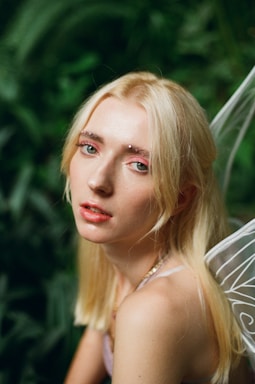 A person with long blonde hair and subtle makeup is looking towards the camera. They are adorned with sparkling decorations on the eyebrows, and they appear to have delicate fairy wings on their back. The background is filled with lush green foliage, giving a natural, woodland ambiance.