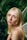 A person with long blonde hair and subtle makeup is looking towards the camera. They are adorned with sparkling decorations on the eyebrows, and they appear to have delicate fairy wings on their back. The background is filled with lush green foliage, giving a natural, woodland ambiance.