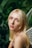 A person with long blonde hair and subtle makeup is looking towards the camera. They are adorned with sparkling decorations on the eyebrows, and they appear to have delicate fairy wings on their back. The background is filled with lush green foliage, giving a natural, woodland ambiance.