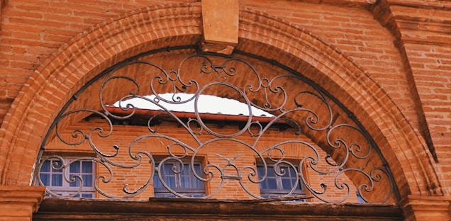 An ornate wrought iron grille with intricate patterns decorates the top of an arched opening. Behind the arch, there is a building made of bricks with two window frames visible. The windows have a slightly reflective glass, and the brickwork is uniform with a reddish hue.