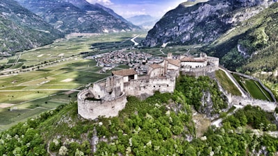 An aerial view of a historic castle nestled on a lush green hilltop, surrounded by agricultural fields and a small village. Towering mountains form a stunning backdrop, enhancing the sense of seclusion and grandeur.