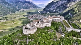 An aerial view of a historic castle nestled on a lush green hilltop, surrounded by agricultural fields and a small village. Towering mountains form a stunning backdrop, enhancing the sense of seclusion and grandeur.