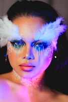A person with dramatic makeup featuring elaborate white feather designs above the eyes, coupled with vibrant blue and pink lighting that highlights the contours of the face. The makeup includes blue eyeshadow and face paint creating a striking look.