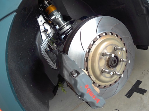 A close-up view of a car's braking system, featuring a large, ventilated brake disc with a high-performance brake caliper labeled 'Brembo'. The suspension components are visible, indicating advanced engineering. The metallic surfaces reflect light, suggesting new or well-maintained parts.