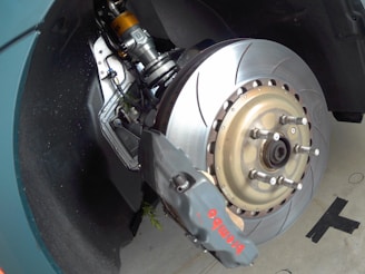 A close-up view of a car's braking system, featuring a large, ventilated brake disc with a high-performance brake caliper labeled 'Brembo'. The suspension components are visible, indicating advanced engineering. The metallic surfaces reflect light, suggesting new or well-maintained parts.