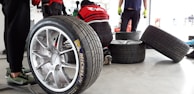 Several racing tires are placed on a concrete floor, with one large tire in the foreground. A person is kneeling and working on a tire, while another person stands nearby wearing green gloves. The background shows a garage environment with tools and equipment.
