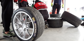 Several racing tires are placed on a concrete floor, with one large tire in the foreground. A person is kneeling and working on a tire, while another person stands nearby wearing green gloves. The background shows a garage environment with tools and equipment.