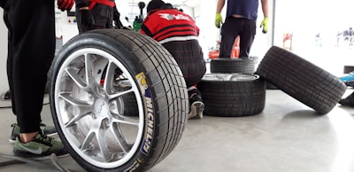 Several racing tires are placed on a concrete floor, with one large tire in the foreground. A person is kneeling and working on a tire, while another person stands nearby wearing green gloves. The background shows a garage environment with tools and equipment.