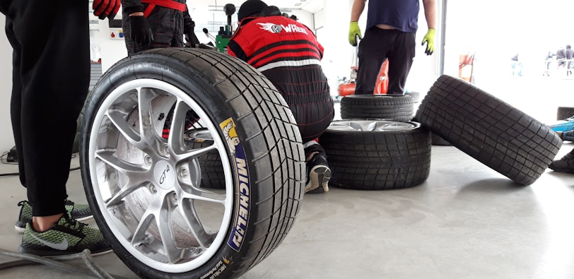 Several racing tires are placed on a concrete floor, with one large tire in the foreground. A person is kneeling and working on a tire, while another person stands nearby wearing green gloves. The background shows a garage environment with tools and equipment.