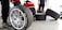 Several racing tires are placed on a concrete floor, with one large tire in the foreground. A person is kneeling and working on a tire, while another person stands nearby wearing green gloves. The background shows a garage environment with tools and equipment.