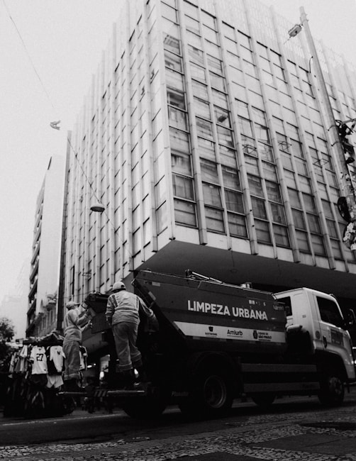 A large urban building dominates the background with a grid of windows, characteristic of mid-20th-century architecture. In the foreground, two sanitation workers in uniform are standing on the back of a garbage truck labeled 'LIMPEZA URBANA.' The scene suggests a city street setting, with utility lines and a street vendor's clothing rack visible.