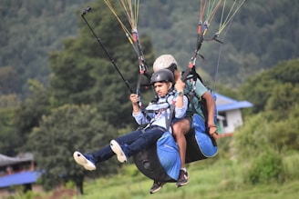 Two people are tandem paragliding, wearing helmets and harnesses, with the landscape and a building visible in the background. They appear to be in mid-air, and one of them is holding a selfie stick.