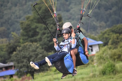 Two people are tandem paragliding, wearing helmets and harnesses, with the landscape and a building visible in the background. They appear to be in mid-air, and one of them is holding a selfie stick.