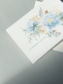 A greeting card featuring a watercolor design of flowers, predominantly in shades of blue and green, with touches of pink and yellow. The card reads 'So grateful for you' and is placed on a neutral-colored surface.