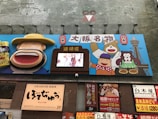 A vibrant billboard with colorful cartoon characters, including a smiling figure wearing a large hat and other animated figures holding traditional Japanese foods. A digital screen in the center displays an image of people. Below the billboard is signage for a restaurant with text in Japanese characters.