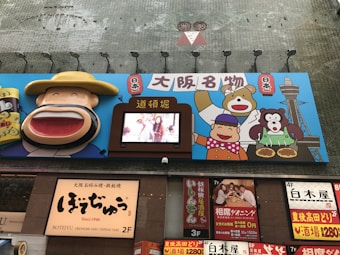 A vibrant billboard with colorful cartoon characters, including a smiling figure wearing a large hat and other animated figures holding traditional Japanese foods. A digital screen in the center displays an image of people. Below the billboard is signage for a restaurant with text in Japanese characters.