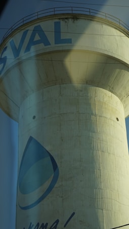 A large cylindrical water tower with a logo and some text partially visible at the top. The surface appears weathered with a pale, off-white color. Some blue graphics are printed on its side. A railing at the top suggests an accessible platform.