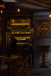 A dimly lit café entrance with warm lighting and a visible bar area. Bottles are arranged on shelves behind the counter, and a few tables and chairs are set outside. The café sign is prominently displayed on the right.