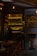 A dimly lit café entrance with warm lighting and a visible bar area. Bottles are arranged on shelves behind the counter, and a few tables and chairs are set outside. The café sign is prominently displayed on the right.