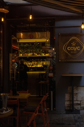 A dimly lit café entrance with warm lighting and a visible bar area. Bottles are arranged on shelves behind the counter, and a few tables and chairs are set outside. The café sign is prominently displayed on the right.