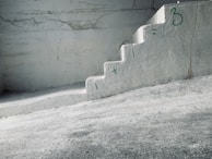 A concrete staircase is painted with a simple math equation in green: '1 + 1 = 3'. The walls and steps have a rough texture with visible cracks and some discoloration. The overall setting has an industrial and minimalistic appearance.