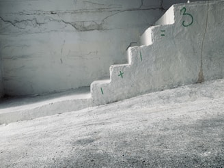 A concrete staircase is painted with a simple math equation in green: '1 + 1 = 3'. The walls and steps have a rough texture with visible cracks and some discoloration. The overall setting has an industrial and minimalistic appearance.