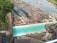 A large dam with turquoise water flowing over it, surrounded by rocky terrain and lush green trees on one side. The water downstream turns into a rocky riverbed. There are red railings and industrial structures visible around the dam.
