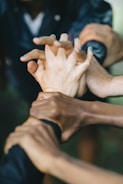 A group of hands interlocked, symbolizing unity and teamwork. The hands are of different skin tones, indicating diversity. The background is slightly blurred, emphasizing the focus on the hands in the center.