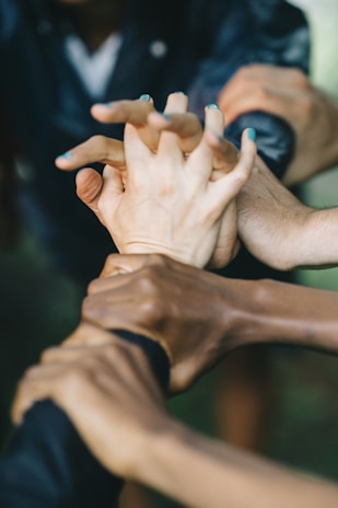 A group of hands interlocked, symbolizing unity and teamwork. The hands are of different skin tones, indicating diversity. The background is slightly blurred, emphasizing the focus on the hands in the center.
