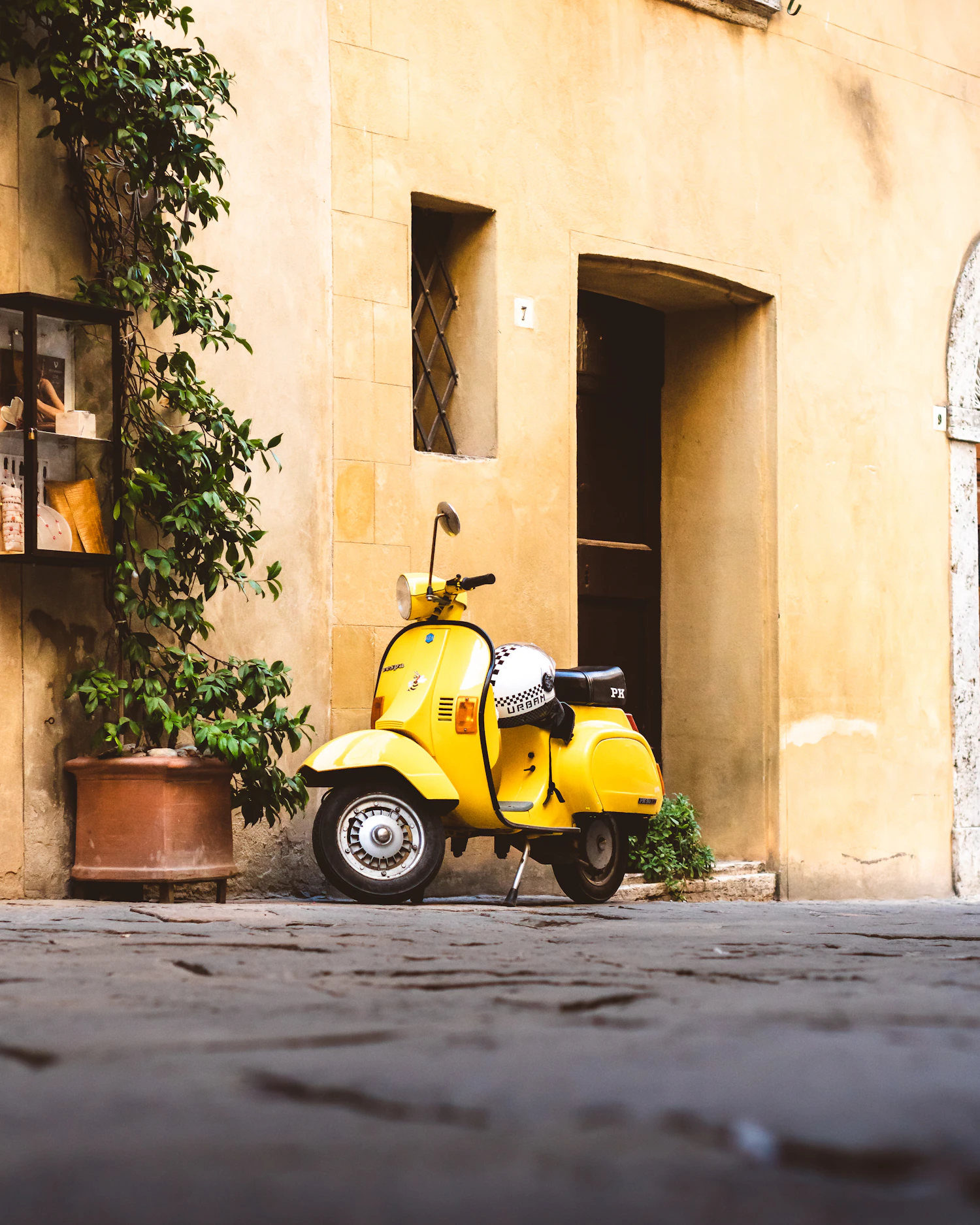 A yellow scooter parked on cobbles against a warm ochre wall, beside a doorway and potted plants