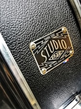 A close-up of a textured black surface with a metallic label featuring the words 'Studio' and design elements like lightning bolts. The label has a glossy finish and is attached to the surface with four screws.