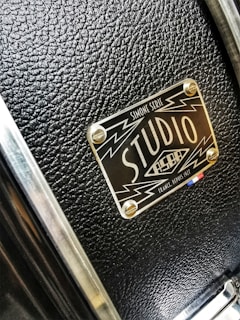 A close-up of a textured black surface with a metallic label featuring the words 'Studio' and design elements like lightning bolts. The label has a glossy finish and is attached to the surface with four screws.