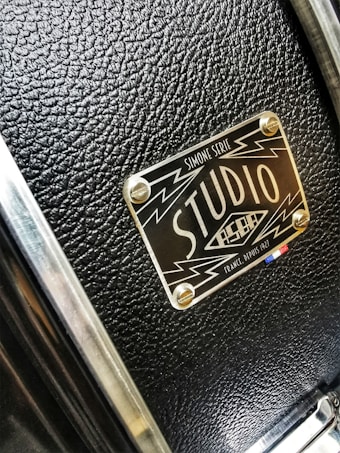 A close-up of a textured black surface with a metallic label featuring the words 'Studio' and design elements like lightning bolts. The label has a glossy finish and is attached to the surface with four screws.