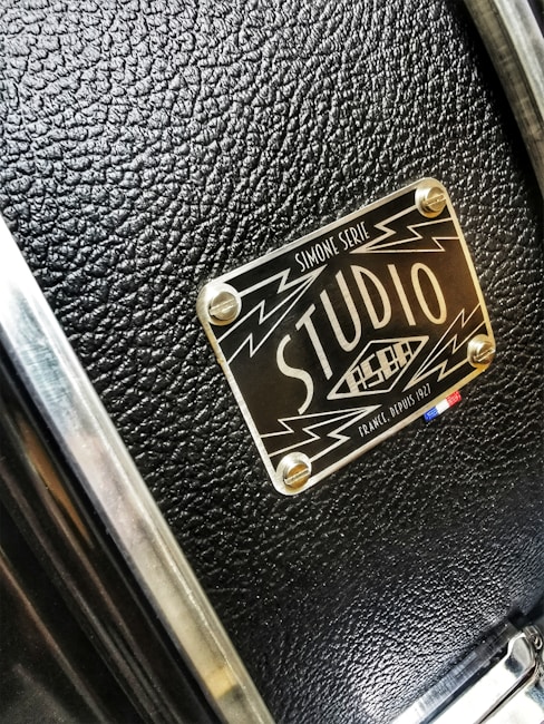 A close-up of a textured black surface with a metallic label featuring the words 'Studio' and design elements like lightning bolts. The label has a glossy finish and is attached to the surface with four screws.