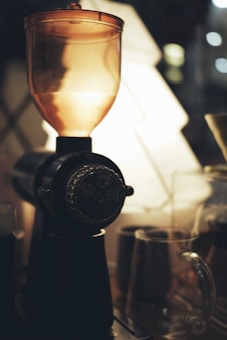 A close-up view of a coffee grinder with a transparent hopper, set against a softly lit background. The focus is on the grinder's central part, with blurred mugs and possibly other coffee-making equipment visible. The lighting creates a warm, cozy atmosphere.
