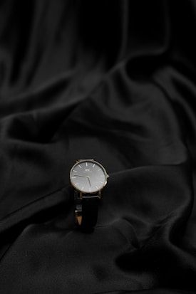 A sleek wristwatch is elegantly placed on a smooth, black fabric. The watch features a simple, minimalist design with a black strap and a silver watch face displaying thin hour markers and hands.