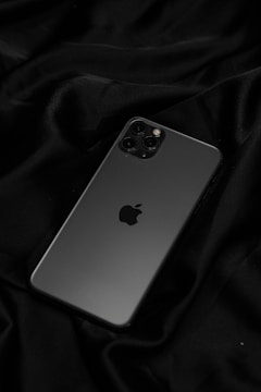 A sleek, dark grey smartphone lies on a black fabric background, showcasing its minimalist design with prominent camera lenses and a logo on the back.
