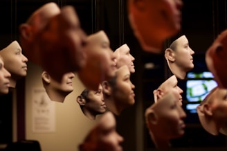 A collection of suspended, realistic human face masks hanging closely together. The masks appear to be made of a lifelike material and vary in skin tones. The background is dark, with some out-of-focus elements such as a screen on the right and a poster on the left.