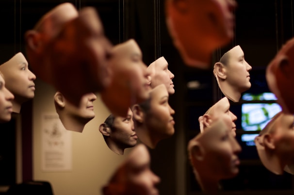A collection of suspended, realistic human face masks hanging closely together. The masks appear to be made of a lifelike material and vary in skin tones. The background is dark, with some out-of-focus elements such as a screen on the right and a poster on the left.